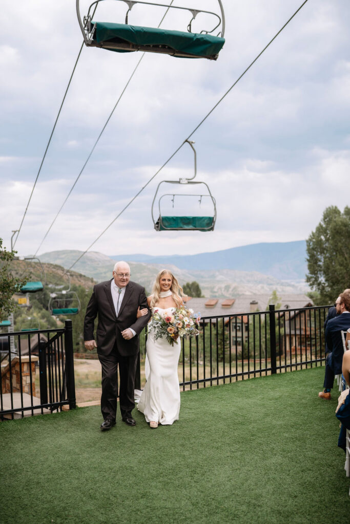 Bride and father walk to ceremony with ski lifts